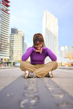 A trendy young woman sits on a city road, vibrant buildings behind herの写真素材