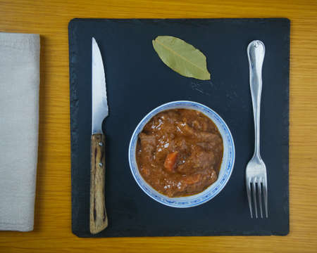 Fricando, Traditional Catalan tapa, stew of beef in a bowl decorated with metal cutlery, a napkin and a bay leaf on a black slate board. Top viewの写真素材