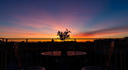 A silhouetted plant on a balcony table overlooks a cityscape during a vibrant orange, purple,...