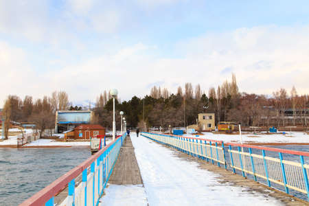 Winter beach. Blue clear sea water, snow and a wooden pier. Skyline and mountains. Kyrgyzstan,...