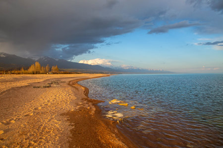 Sunset on the shore of Lake Issyk-Kul, Kyrgyzstan, with soft sandy beaches and a cloudy sky against the mountainsの写真素材