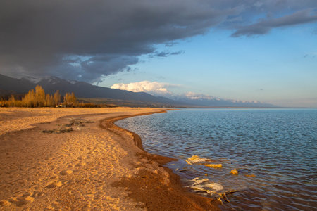 Sunset on the shore of Lake Issyk-Kul, Kyrgyzstan, with soft sandy beaches and a cloudy sky against the mountainsの写真素材