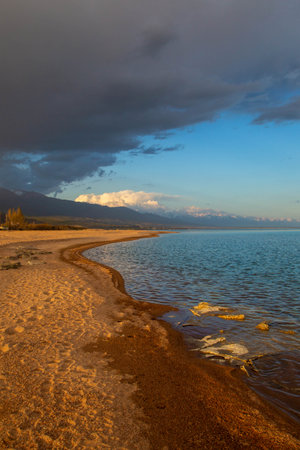 Sunset on the shore of Lake Issyk-Kul, Kyrgyzstan, with soft sandy beaches and a cloudy sky against the mountainsの写真素材