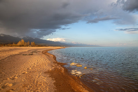 Sunset on the shore of Lake Issyk-Kul, Kyrgyzstan, with sandy beaches and a cloudy sky against the mountainsの写真素材