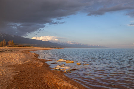 Sunset on the shore of Lake Issyk-Kul, Kyrgyzstan, with soft sandy beaches and a cloudy sky against the mountainsの写真素材