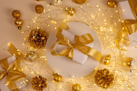 Golden lights, pine cones and white gift with ribbon on warm beige background. Minimal and festive holiday composition.の写真素材
