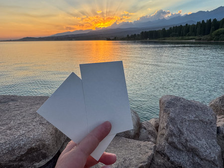 Two blank business cards held in hand against scenic sunset over lake and mountains. Natural atmospheric branding mockup outdoors.の写真素材