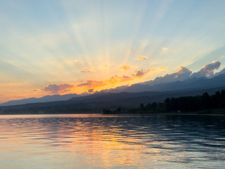 Sunset at Issyk Kul Lake in Kyrgyzstan. Sun rays breaking through clouds reflecting in calm water with mountains in background. Peaceful sunset at Issyk Kul with sun reflection and Tien Shan mountains.の写真素材