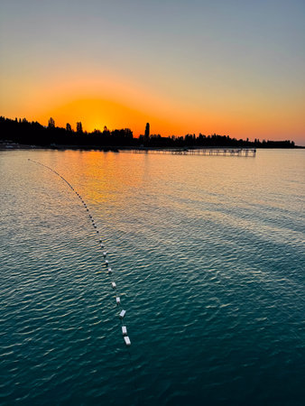 Early sunrise at Issyk Kul Lake in Kyrgyzstan. Warm golden sky reflecting in calm water with silhouettes of trees and pier on horizon. Sunrise at Issyk Kul with golden horizon and calm water surface.の写真素材