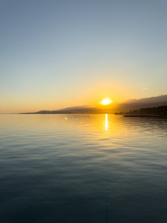 Scenic sunset over Issyk Kul Lake in Kyrgyzstan. The sun sets behind the mountains, casting a golden reflection across the water. Sunset over Issyk Kul Lake in Kyrgyzstan with golden reflection on the water.の写真素材