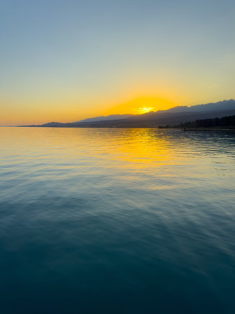 Scenic sunset over Issyk Kul Lake in Kyrgyzstan. The sun sets behind the mountains, casting a golden reflection across the water. Sunset over Issyk Kul Lake in Kyrgyzstan with golden reflection on the water.の写真素材