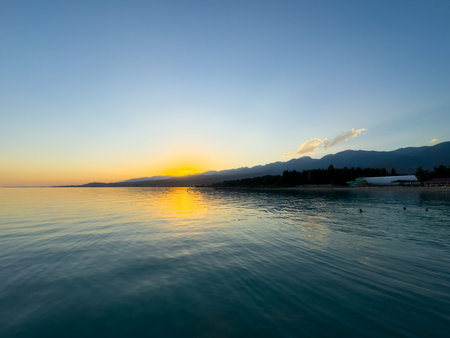Scenic sunset over Issyk Kul Lake in Kyrgyzstan. The sun sets behind the mountains, casting a golden reflection across the water. Sunset over Issyk Kul Lake in Kyrgyzstan with golden reflection on the water.の写真素材