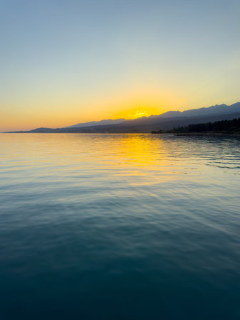 Scenic sunset over Issyk Kul Lake in Kyrgyzstan. The sun sets behind the mountains, casting a golden reflection across the water. Sunset over Issyk Kul Lake in Kyrgyzstan with golden reflection on the water.の写真素材