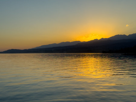 Scenic sunset over Issyk Kul Lake in Kyrgyzstan. The sun sets behind the mountains, casting a golden reflection across the water. Sunset over Issyk Kul Lake in Kyrgyzstan with golden reflection on the water.の写真素材