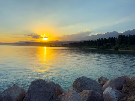 Scenic sunset over Issyk Kul Lake in Kyrgyzstan. The sun sets behind the mountains, casting a golden reflection across the water. Sunset over Issyk Kul Lake in Kyrgyzstan with golden reflection on the water.の写真素材