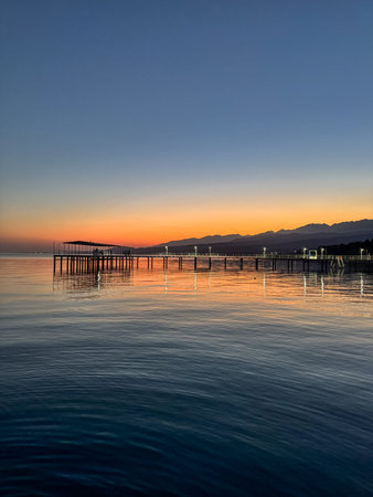 Evening sunset at Issyk Kul Lake in Kyrgyzstan. Long illuminated pier stretching into calm water with mountains on horizon. Sunset at Issyk Kul with long pier and light reflections on calm water.の写真素材
