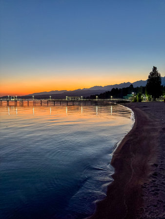 Evening sunset with long illuminated pier stretching into calm water with mountains on horizon. Sunset with long pier and light reflections on calm water.の写真素材