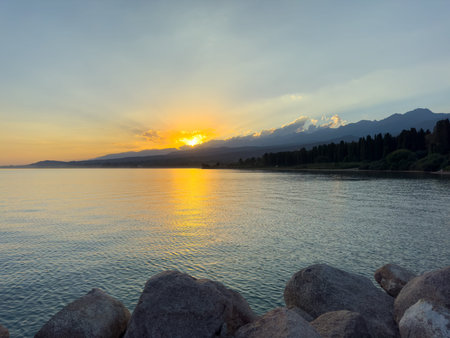 Scenic sunset over Issyk Kul Lake in Kyrgyzstan. The sun sets behind the mountains, casting a golden reflection across the water. Sunset over Issyk Kul Lake in Kyrgyzstan with golden reflection on the water.の写真素材