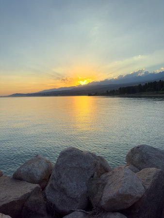 Scenic sunset over Issyk Kul Lake in Kyrgyzstan. The sun sets behind the mountains, casting a golden reflection across the water. Sunset over Issyk Kul Lake in Kyrgyzstan with golden reflection on the water.の写真素材