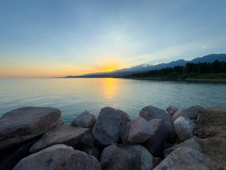 Scenic sunset over Issyk Kul Lake in Kyrgyzstan. The sun sets behind the mountains, casting a golden reflection across the water. Sunset over Issyk Kul Lake in Kyrgyzstan with golden reflection on the water.の写真素材