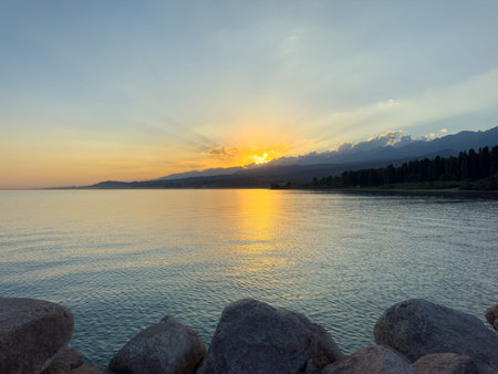 Scenic sunset over Issyk Kul Lake in Kyrgyzstan. The sun sets behind the mountains, casting a golden reflection across the water. Sunset over Issyk Kul Lake in Kyrgyzstan with golden reflection on the water.の写真素材