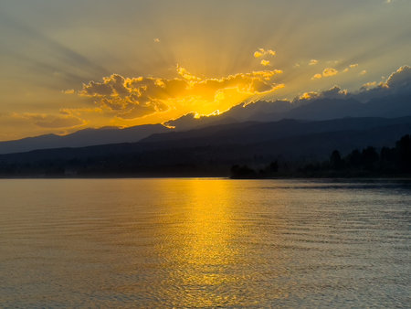 Scenic sunset over Issyk Kul Lake in Kyrgyzstan. The sun sets behind the mountains, casting a golden reflection across the water. Sunset over Issyk Kul Lake in Kyrgyzstan with golden reflection on the water.の写真素材