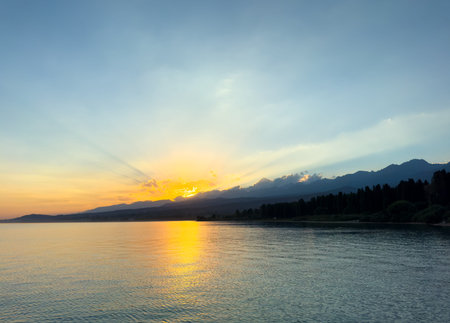 Scenic sunset over Issyk Kul Lake in Kyrgyzstan. The sun sets behind the mountains, casting a golden reflection across the water. Sunset over Issyk Kul Lake in Kyrgyzstan with golden reflection on the water.の写真素材