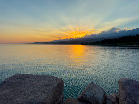 Scenic sunset over Issyk Kul Lake in Kyrgyzstan. The sun sets behind the mountains, casting a golden reflection across the water. Sunset over Issyk Kul Lake in Kyrgyzstan with golden reflection on the water.の写真素材