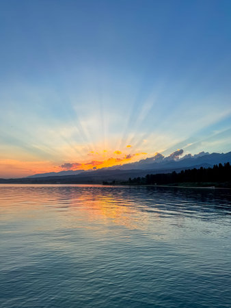 Sunset at Issyk Kul Lake in Kyrgyzstan. Sun rays breaking through clouds reflecting in calm water with mountains in background. Peaceful sunset at Issyk Kul with sun reflection and Tien Shan mountains.の写真素材