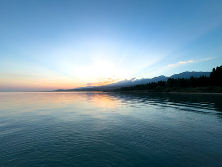 Sunset at Issyk Kul Lake in Kyrgyzstan. Sun rays breaking through clouds reflecting in calm water with mountains in background. Peaceful sunset at Issyk Kul with sun reflection and Tien Shan mountains.の写真素材
