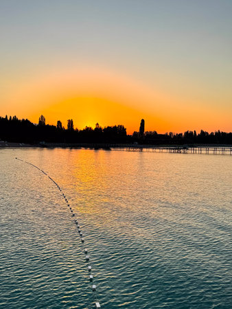 Early sunrise at Issyk Kul Lake in Kyrgyzstan. Warm golden sky reflecting in calm water with silhouettes of trees and pier on horizon. Sunrise at Issyk Kul with golden horizon and calm water surface.の写真素材