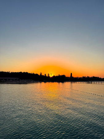 Early sunrise at a lake. Warm golden sky reflecting in calm water with silhouettes of trees and pier on horizon. Sunrise with golden horizon and calm water surface.の写真素材