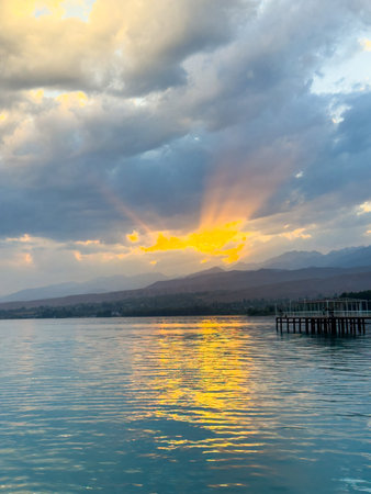 Sunset at Issyk Kul Lake in Kyrgyzstan. Bright sun breaking through clouds reflecting in clear turquoise water. Turquoise water of Issyk Kul at sunset with golden reflection and dramatic clouds.の写真素材