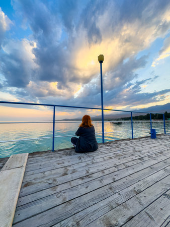 Issyk Kul Lake, Kyrgyzstan. A woman sits on a wooden pier watching the sunset sky reflected in calm water. Woman sitting on a pier watching sunset over Issyk Kul Lake in Kyrgyzstan.の写真素材