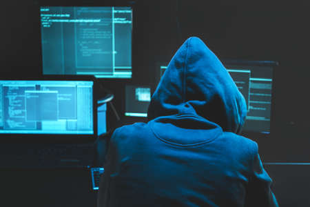 Hacker using computer for organizing massive data breach attack on goverment servers. Hacker in dark room surrounded computersの写真素材