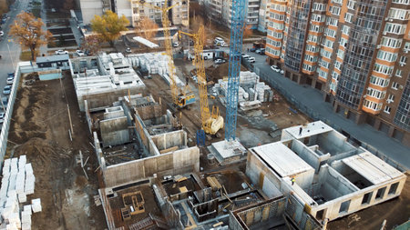 An aerial view of a construction site in a city. A large crane dominates the center of the frame,...