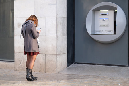 Urban dweller in stylish attire engrossed in smartphone near an outdoor ATM. Woman at Bank using smartphone.の写真素材
