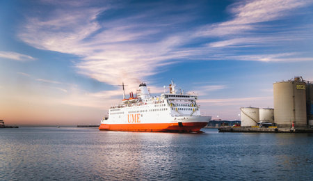 Large orange and white ferry boat anchored at port during sunset, with industrial structures visible...