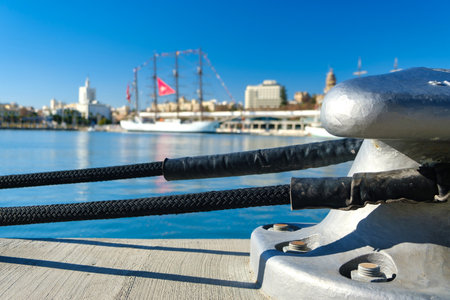 Close-up of metallic bollard with black ropes at dock under blue sky, with distant bridge and cityscape viewsの写真素材