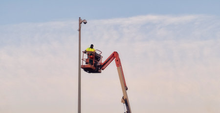 Worker in safety gear operating at height on lift, servicing pole equipment against clear sky backdropの写真素材