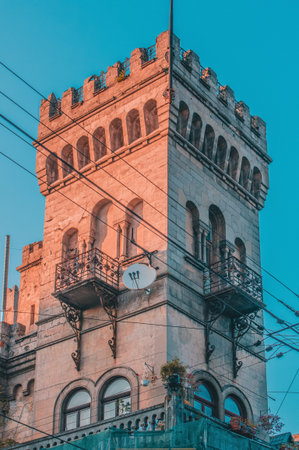The image shows a tall building with architectural features resembling a medieval tower or small castle. It has a rectangular shape with battlements at the top, reminiscent of a crenellated parapet that would be used for defense in medieval times. There are multiple levels with arched windows, and some have balconies with intricate iron railings. Plants can be seen on one of the balconies, adding a touch of greenery to the structure. The building appears to be situated in an urban area, as suggested by the presence of electrical wires in front of it. The lighting suggests either dawn or dusk, giving the picture a warm tone.の写真素材