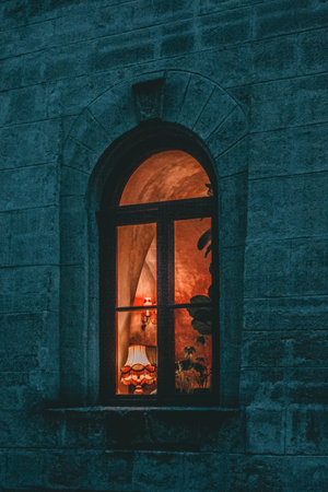 The image shows an arched window set into a stone wall during twilight or night time. The window is lit from the inside, casting a warm glow. Through the pane, one can see indoor plants and what looks like a lamp with a decorative shade, which contributes to the cozy atmosphere. The image has a moody aesthetic, achieved through the contrast between the warm interior light and the cool blue tones of the exterior. It evokes a feeling of warmth and solitude.の写真素材