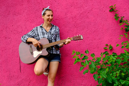 Young woman playing acoustic guitar on pink wall and green leaves background. Hipster girl portrait...