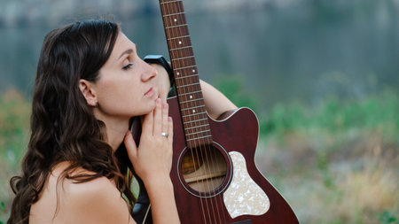 Attractive long haired, looking at the her guitar. Outdoor portrait with nature background, a young and beautiful model. Portrait of young woman with a guitarの写真素材