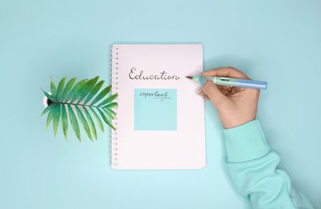 Female hand holding a pen and writing education in notepad. Flat lay on turquoise backgroundの写真素材