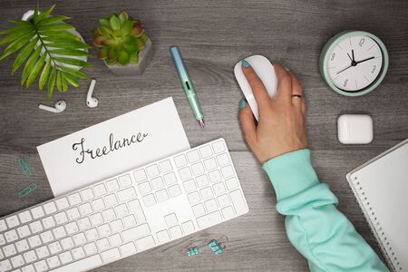 Female hand holding a mouse. Word freelance in notepad. Business flat layout with palm and plant on gray wooden backgroundの写真素材