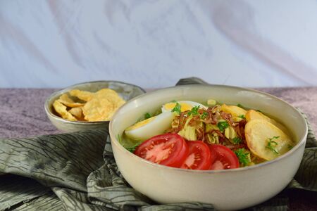 Soto Ayam, yellow turmeric chicken soup with vermicelli noodles, shredded chicken, boiled egg, fried potato, sprouts, tomato, parsley and fried shallots, served with emping. Popular Indonesian foodの写真素材