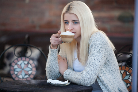 A blonde woman sits in restaurant, drinking coffee and looking through window. Bright and reflection sceneの写真素材