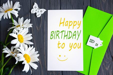 Happy birthday lettering with camomile flowers and a card on a dark wooden background. Greeting card for the birthday of mom, daughter, girl.