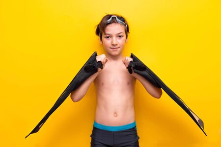 Travel and vacation. Close-up of a 9 years old boy with goggles for swimming on his head with swimming flippers in his hands. Yellow background.の写真素材